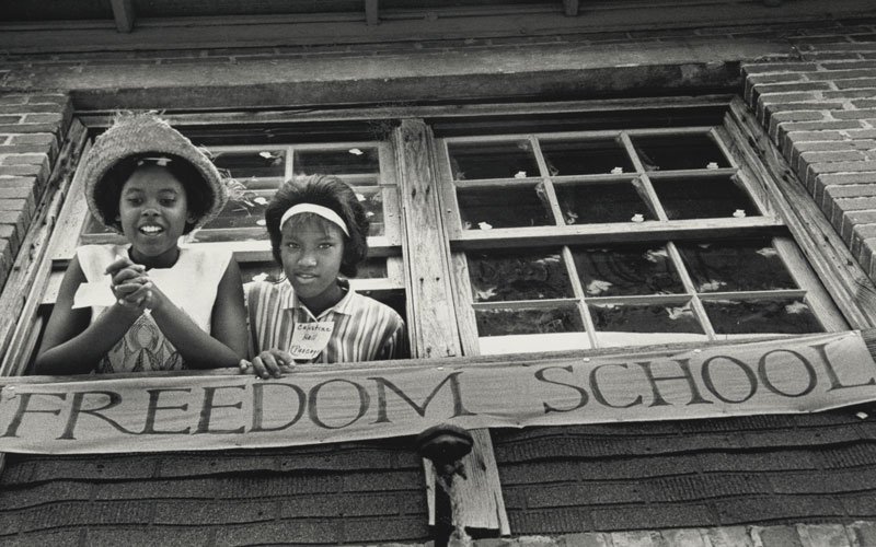 Students leaned out of the window smiling above a banner that says Freedom School at 1964 Freedom School in Mississippi